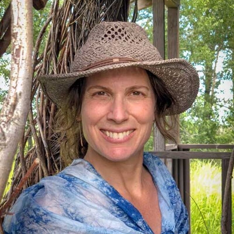 The image shows a woman with fair skin, wearing a woven hat and a blue and white patterned shawl. She has shoulder-length brown hair and is smiling at the camera. The background includes trees and some wooden structures, suggesting an outdoor setting. The lighting is natural and soft, highlighting her features.